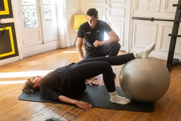 Un homme observe une femme faire un pont sur un tapis, les pieds sur un ballon de gym. Un exercice pour le bienfait d'étirement et de renforcement.